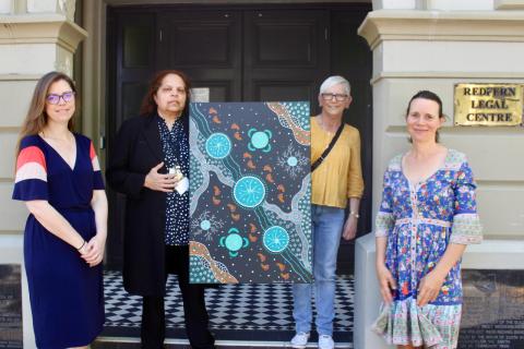 Four women stand outside a building, the two in the centre hold up a green plack and organing ABorigial artwork, a gold plaque behind the woman furtherest to the right reads "Redfern Legal Centre"